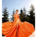 A woman stands outdoors on a wooden deck, wearing the SIENNA NC2052 gown, which features a vibrant sienna hue with a flowing skirt and high slit. The sunlight casts a warm glow as tall evergreen trees frame the scene. She poses gracefully, holding out the sides of her dress. Angels Formal Wear