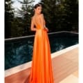 A woman stands beside a pool, facing away from the camera, wearing an elegant, sleeveless, backless SIENNA NC2052 orange gown that drapes to the floor. She is surrounded by tall evergreen trees under a clear sky. Angels Formal Wear