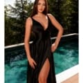 A woman with long brown hair stands by a pool, surrounded by greenery. She is wearing a sleeveless black SIENNA NC2052 gown with a deep neckline and a high slit on one side; the elegant attire epitomizes the luxurious style of the SIENNA collection. With one hand on her hip, she looks directly at the camera. Angels Formal Wear