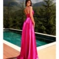 A woman in a flowing, bright pink dress stands by an outdoor pool overlooking lush green mountains. Her back is turned towards the camera as she gazes over her shoulder, exuding an air of elegance. This picturesque scene, reminiscent of a SIENNA NC2052 postcard, features vibrant colors and a serene, natural backdrop. Angels Formal Wear