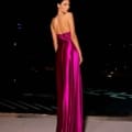 A woman is standing on an outdoor terrace at night, looking over her shoulder. She is wearing the NATALIA NC2009, a floor-length, backless, vibrant magenta satin gown. The dark sky and dimly lit background create a striking contrast with her bright dress. Angels Formal Wear