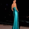 A woman in the sophisticated NATALIA NC2009 evening gown poses with her back to the camera. This floor-length, strapless teal dress features a shiny satin finish. The dark background accentuates the gown's vibrant color and showcases her poised, graceful stance. Angels Formal Wear