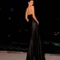 A woman stands elegantly at night, adorned in the sleek, strapless NATALIA NC2009 evening gown. She faces away from the camera, her hands resting in the gown's pockets, which accentuate its long and flowing design. The dimly lit background enhances her silhouette beautifully. Angels Formal Wear