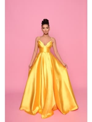 Against a pink backdrop, a woman elegantly poses in a striking floor-length BRENNA NP1 gown. The vibrant yellow dress features thin straps and a deep neckline. She holds the sides of the gown, slightly spreading it to display its fullness, while her hair is styled in an elegant updo. Angels Formal Wear