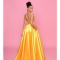 A woman stands against a pink background, facing slightly away from the camera. She is wearing the BRENNA NP157, a long, yellow satin gown with thin straps and an open back. Her hair is styled in an elegant updo, and she gently touches the side of her dress with one hand, embodying the grace of BRENNA's latest collection. Angels Formal Wear