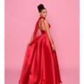 A woman poses elegantly against a pink backdrop, wearing a striking, floor-length red satin gown with a flowing train. Her hair is styled in an intricate updo, and she wears dangling earrings. The confident expression on her face captures the essence of BRENNA NP157 effortlessly. Angels Formal Wear