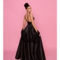 Against a solid pink background, a woman poses gracefully, wearing the elegant long BRENNA NP157 black gown with thin straps. Her hair is styled in an intricate updo, and as she faces slightly away from the camera, she showcases the gown's open back design. Her poise evokes timeless beauty in this exquisite attire. Angels Formal Wear