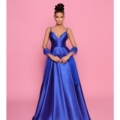 Brenna, wearing the BRENNA NP157, a floor-length, royal blue satin gown, stands against a pink background. The dress features spaghetti straps, a fitted bodice, and a full skirt. She holds the hem of the skirt with both hands; her hair is styled in an elegant updo. Angels Formal Wear