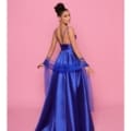 A woman in a glamorous, royal blue satin evening gown from BRENNA's NP157 collection poses against a pink background. The dress features thin straps and a fitted bodice with a flowing skirt. She holds a matching sheer blue shawl, and her dark hair is styled in an elegant updo. Angels Formal Wear