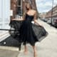 A woman with long hair poses on a city sidewalk wearing a strapless black dress and beige high heels, embodying the elegance of Product JX6047. She holds the sides of her dress, creating a flowing effect. The background features a row of brick buildings, white doors, and a partly cloudy sky. Angels Formal Wear