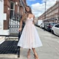 A woman wearing the JADORE JX6047 strapless white dress, looking effortlessly chic, stands on a sidewalk in an urban street lined with brick buildings and parked cars. Leaning against a wrought iron fence, she glances to the side. The sky is partly cloudy—JADORE captures this moment with timeless elegance. Angels Formal Wear