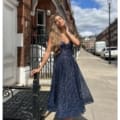 A woman stands on a city sidewalk, gracefully leaning against a black iron railing. She is elegantly dressed in the sleeveless, ankle-length JADORE JX6047 navy blue dress, adorned with sparkling details. Behind her are red brick buildings, a clear sky, and a street lamp. Angels Formal Wear