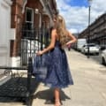 A woman with long, wavy blonde hair stands on a city sidewalk with her back to the camera. She wears the JADORE JX6047, a sparkly dark blue dress paired with high-heeled sandals, exuding an effortlessly chic vibe. Row houses with red brick facades line the street, and it's a sunny day with scattered clouds against the blue sky. Angels Formal Wear