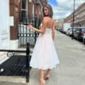 A woman with long hair, wearing a white, strapless JADORE JX6047 knee-length dress and high-heeled shoes, walks on a sidewalk in an urban area. She looks over her shoulder towards the camera. The background includes a row of brick buildings, parked cars, and a blue sky. Angels Formal Wear