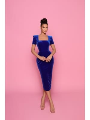 Against a pink backdrop, a woman exudes confidence in the stunning JADORE NP160 dress, a form-fitting masterpiece in royal blue velvet. With short sleeves and a square neckline, she complements the look with her hair elegantly styled up and nude heels, epitomizing sophistication. Angels Formal Wear