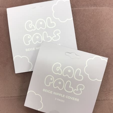 Two packages of "GAL PALS NIPPLE COVERS," each containing five pairs, rest on a soft brown quilted surface. The charming cloud-designed packaging adds a playful touch to these delightful beige nipple shields. Angels Formal Wear