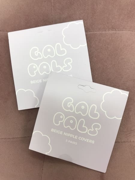 Two packages of "GAL PALS NIPPLE COVERS," each containing five pairs, rest on a soft brown quilted surface. The charming cloud-designed packaging adds a playful touch to these delightful beige nipple shields. Angels Formal Wear