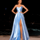 A woman wearing a strapless, light blue satin gown with a thigh-high slit stands confidently with her hands on her hips. Her long hair is styled straight as she poses outdoors at night by the HAVANNA NC2030 swimming pool, with soft evening lighting enhancing the luxurious atmosphere. Angels Formal Wear