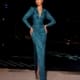 A woman wearing the FOXETTE NC2011, a long-sleeved, sparkling teal gown with a deep V-neckline and a thigh-high slit, stands on a tiled floor at night. Her dress reveals silver high heels beneath the slit. The background is dark with distant lights, evoking the glamorous NC2011 era. Angels Formal Wear