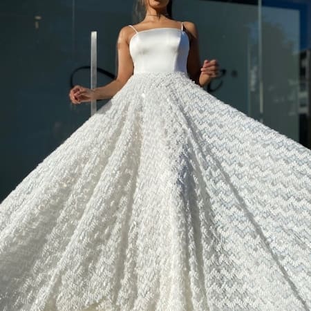 A woman wearing the BRIAR JX4056 white sleeveless gown poses confidently outdoors. The dress features a fitted bodice and a voluminous textured skirt with a wavy pattern. She stands in front of a glass building, with sunlight highlighting her attire. Angels Formal Wear