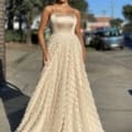 A woman stands outdoors wearing a long, elegant BRIAR JX4056 beige gown with a fitted bodice and a voluminous textured skirt. She has long, straight hair, and the background includes trees, a street, and parked cars. The sunlight highlights her dress and figure beautifully. Angels Formal Wear