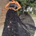 A woman stands confidently outdoors in a flowing BRIAR JX4056 gown with intricate designs. The dress boasts a fitted bodice and a voluminous skirt adorned with shimmering details. With one hand on her hip, she poses near a brick wall, greenery serving as the backdrop.
 Angels Formal Wear