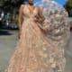 A woman stands outdoors wearing the FLOWERS JX4025, a flowing lace gown adorned with intricate embroidered flowers in shades of beige and peach. The dress features a deep V-neck design. She poses confidently with one hand on her hip against a backdrop that includes a tree and urban elements. Angels Formal Wear