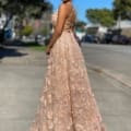 A woman with curly hair is standing outdoors on a city street, adorned in the elegant, floor-length FLOWERS JX4025 beige lace gown featuring a crisscross back design. Amidst blooming flowers, she looks over her shoulder to highlight the stunning back of the dress while sunlight bathes the scene in warmth. Angels Formal Wear