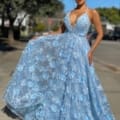 A woman stands outdoors on a sunny day, wearing the floor-length FLOWERS JX4025 dress adorned with blue floral patterns and featuring a deep V neckline. She holds onto the skirt of the dress, slightly lifting it. Trees are visible in the background along with a sidewalk, road, and other urban elements. The FLOWERS JX4025 design accentuates her elegance perfectly. Angels Formal Wear