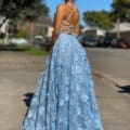 A woman with curly hair, wearing a blue lace FLOWERS JX4025 gown, stands outdoors on a sunny day. The gown features intricate floral patterns and crisscross straps on the back. Flowers bloom in the background alongside trees, cars, and buildings. Angels Formal Wear