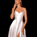 A woman stands elegantly against a black background, touching her face with one hand and looking to the side. She is wearing the HAVANNA NC2030, a white satin strapless gown with a sweetheart neckline and a high slit. The lighting highlights the sheen of her dress and her poised posture. Angels Formal Wear