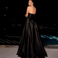 A woman wearing the HAVANNA NC2030 strapless black gown stands by a pool at night. The gown features a fitted bodice and a flowing skirt. Her hair is styled in a bun, and she poses elegantly with her back slightly turned and her arms crossed, gazing over her shoulder. Angels Formal Wear