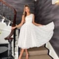 A woman donning a textured white dress stands elegantly on a staircase with an ornate black railing in Brooklyn. She gracefully holds her dress to the side, her long hair gathered in a ponytail. The setting features dark, textured walls and patterned flooring, lending an elegant touch inspired by the BROOKLYN JP125 design. Angels Formal Wear