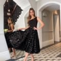 A woman, wearing a chic black sleeveless top and a textured black skirt, poses gracefully in a Brooklyn hallway featuring black and white checkered flooring. Positioned near a large mirror, she complements her look with silver heels. Behind her, the elegant staircase reflects the stylish vibe of the BROOKLYN JP125 collection. Angels Formal Wear