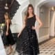 A woman stands in front of a mirror wearing the BROOKLYN JP125, a stylish black dress featuring a ruffled skirt. The room, exuding chic Brooklyn charm, is adorned with elegant decor, showcasing a monochrome tiled floor and an ornate staircase in the background. She poses confidently with one hand on her hip. Angels Formal Wear