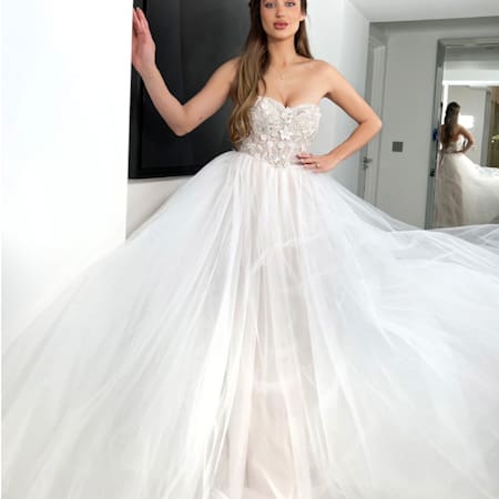 Wearing an EVE JX6006W gown, a woman stands gracefully by a wall with her hand gently resting on it. The strapless dress features intricate floral detailing on the bodice and a voluminous tulle skirt, while a mirror behind her elegantly captures her reflection. Angels Formal Wear