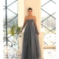 In an elegant room with large windows, a woman stands poised in the stunning gray IVY 1064 strapless floor-length gown. Paired with sheer gloves and her hair styled in a sleek bun, she exudes confidence. Angels Formal Wear