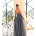 Dressed in the sophisticated, floor-length IVY 1064 gown, a woman stands by an ornate balcony and gazes over her shoulder. In the background, large windows showcase a crisscross design and reveal a hint of the outdoors. Angels Formal Wear