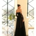 A woman stands indoors in front of large windows wearing an elegant black gown reminiscent of the IVY 1064. The dress features sheer sleeves and a layered skirt. Her hair is styled in an updo as she gazes over her shoulder, while sunlight streams in, casting soft shadows. Angels Formal Wear