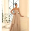 Against a white wall, a woman exudes confidence in the IVY 1064, an elegant gown featuring a strapless sheer beige design with a fitted corset bodice and full skirt. Her hair is styled in an updo as she gracefully poses with one arm resting on a railing. Angels Formal Wear