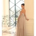 A woman in an elegant strapless gown, labeled as the product IVY 1064, stands gracefully by a decorative railing. She poses sideways, gazing at the camera. The setting boasts large windows and classical architecture, contributing to a sophisticated ambiance. Angels Formal Wear