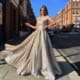 A woman in a shimmering silver gown poses on a city street, embodying the elegance of the BRITTA JX6005 (Copy). Her dress features a flowing, voluminous skirt as she stands confidently before red brick buildings beneath a clear blue sky. Angels Formal Wear