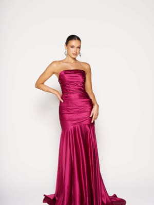 A woman elegantly poses in a strapless satin MOSELLE PO2457 gown in fuchsia, showcasing its fitted bodice and flared hem. She stands against a plain white backdrop, with her hair pulled back and highlighted by large hoop earrings. Angels Formal Wear