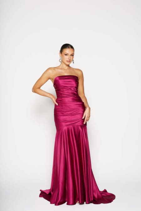 A woman elegantly poses in a strapless satin MOSELLE PO2457 gown in fuchsia, showcasing its fitted bodice and flared hem. She stands against a plain white backdrop, with her hair pulled back and highlighted by large hoop earrings. Angels Formal Wear