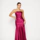 A woman elegantly poses in a strapless satin MOSELLE PO2457 gown in fuchsia, showcasing its fitted bodice and flared hem. She stands against a plain white backdrop, with her hair pulled back and highlighted by large hoop earrings. Angels Formal Wear