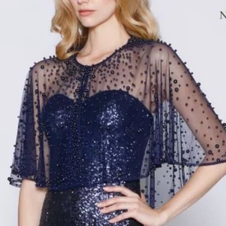 A person wearing a navy blue sequined dress with a sheer, beaded overlay exudes elegance. The overlay features short, flowing sleeves adorned with small round beads. Their light blonde hair is styled in soft waves, impeccably complemented by the stylish Pearl Capes that complete the chic ensemble. Angels Formal Wear