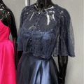 A mannequin elegantly displays the BEADED CAPE, a dark blue piece adorned with intricate beadwork, over a shiny blue gown. Alongside it stands another mannequin in a vibrant pink dress. This clothing arrangement underscores the exquisite craftsmanship of the BEADED CAPE, enhancing the elegance and charm of the ensemble. Angels Formal Wear